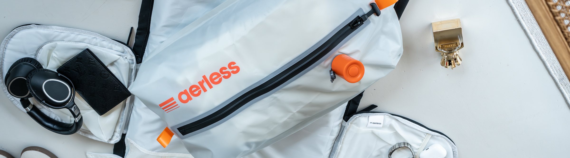 Overhead flat lay of a packed trip: the Aerless bag drawn down flat with its orange pump resting on it, a second bag, over-ear headphones and a wallet laid out around it
